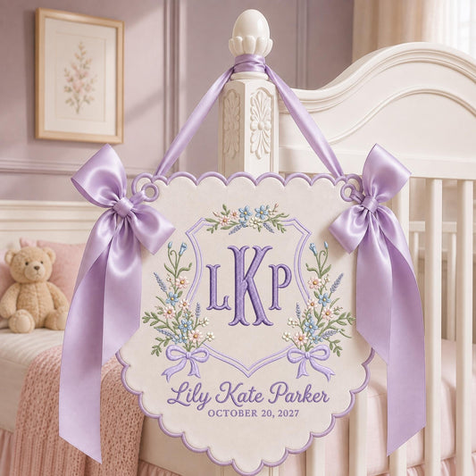 May include: A cream-colored fabric wall hanging with a scalloped edge, featuring purple embroidered monogram letters LKP, floral accents, and the text Lily Kate Parker OCTOBER 20, 2027. It is suspended by a lavender satin ribbon with two large bows.