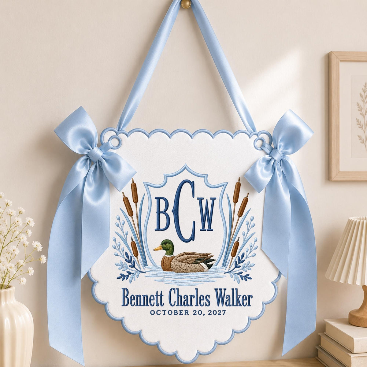 May include: A white scalloped wall hanging featuring a mallard duck illustration, cattails, and a blue embroidered monogram BCW. The text reads Bennett Charles Walker and OCTOBER 20, 2027. It is accented with light blue satin ribbons and bows.