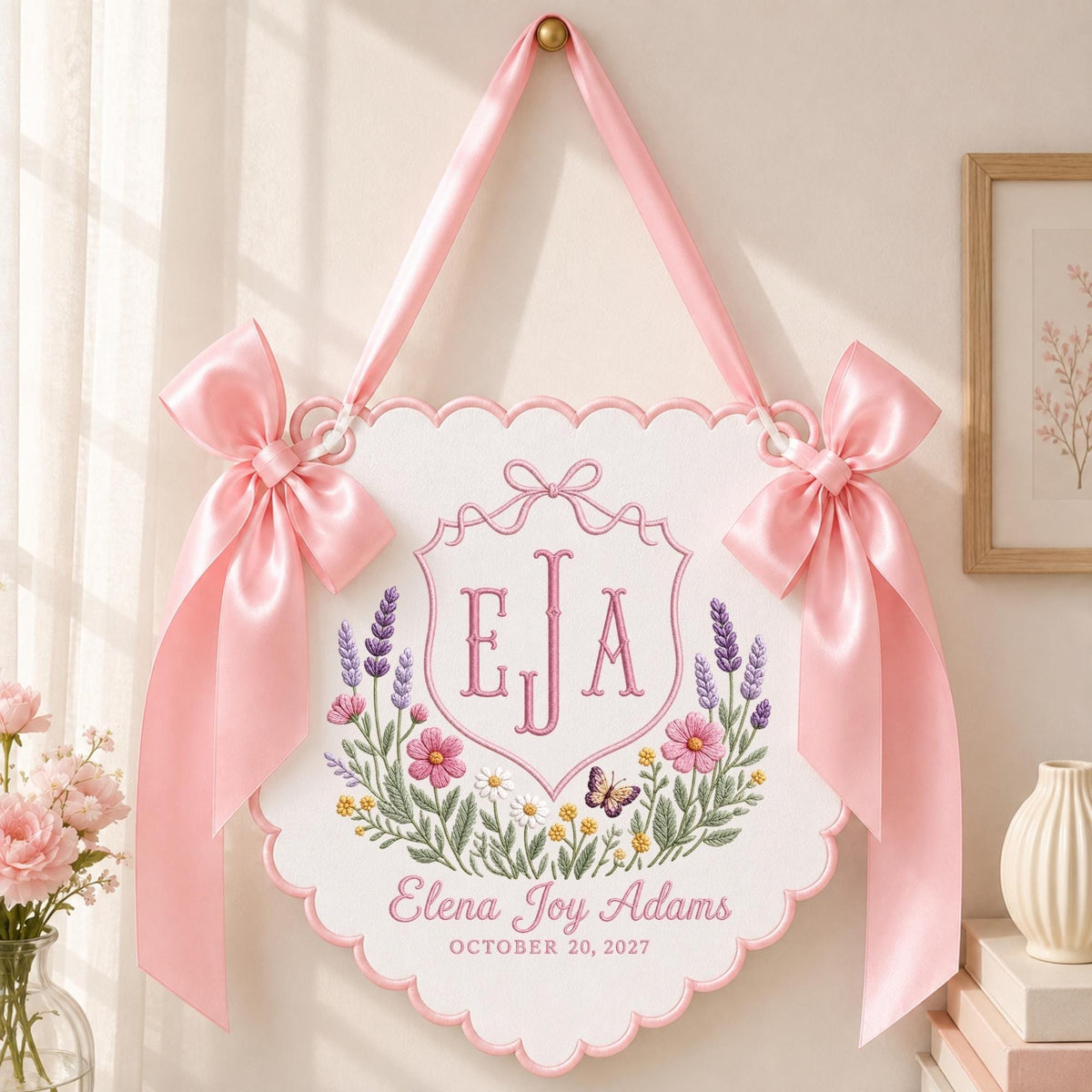 May include: A white scalloped wall hanging featuring pink satin bows, embroidered with the monogram EJA, a floral wreath with lavender, pink, and yellow flowers, a small butterfly, and the text Elena Joy Adams, October 20, 2027.