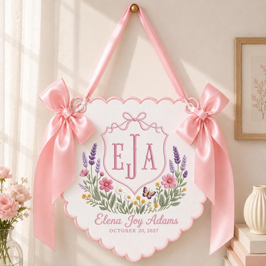 May include: A white scalloped wall hanging featuring pink satin bows, embroidered with the monogram EJA, a floral wreath with lavender, pink, and yellow flowers, a small butterfly, and the text Elena Joy Adams, October 20, 2027.