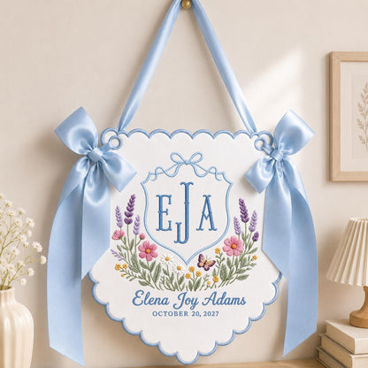 May include: A white scalloped wall hanging featuring blue embroidery of the initials EJA inside a crest, surrounded by lavender, pink, and yellow flowers and a butterfly. The text reads Elena Joy Adams and OCTOBER 20, 2027, with blue satin ribbons.