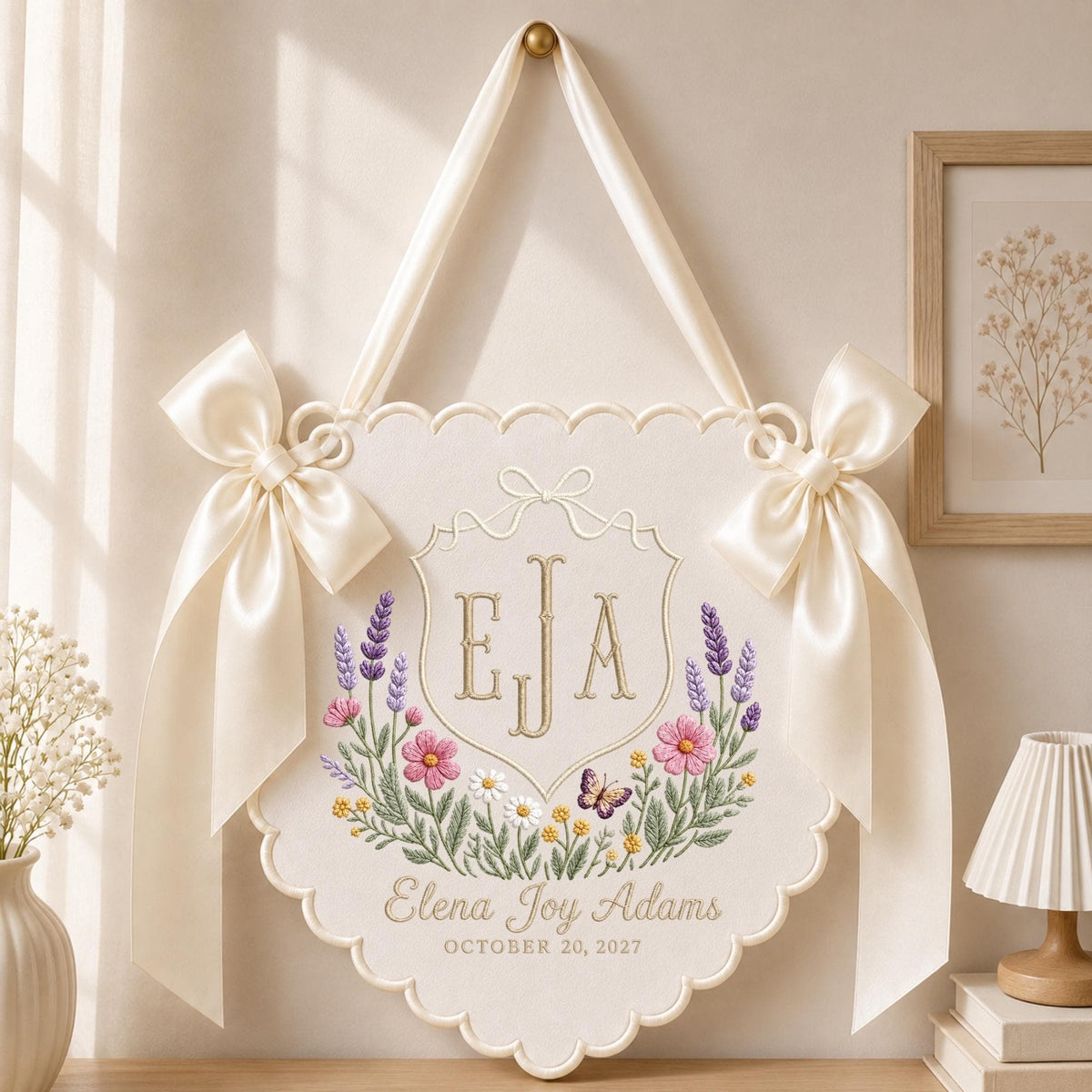 May include: A cream-colored fabric wall hanging with scalloped edges, featuring an embroidered crest with the initials EJA, a floral wreath with lavender and pink flowers, a butterfly, and the text Elena Joy Adams OCTOBER 20, 2027.