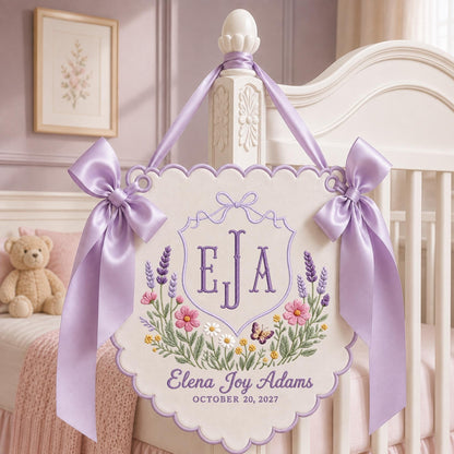 May include: A cream-colored fabric wall hanging with scalloped edges, featuring purple embroidered initials EJA inside a crest, surrounded by floral motifs and a butterfly. It includes the text Elena Joy Adams and October 20, 2027, with lavender satin bows.