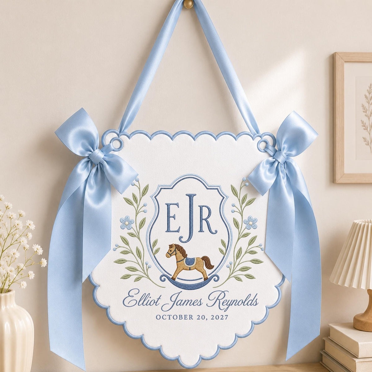 May include: A white scalloped wall hanging banner featuring light blue satin ribbon bows and a blue embroidered crest. The design includes the initials EJR, a brown rocking horse, green leafy branches, and the text Elliot James Reynolds October 20, 2027.