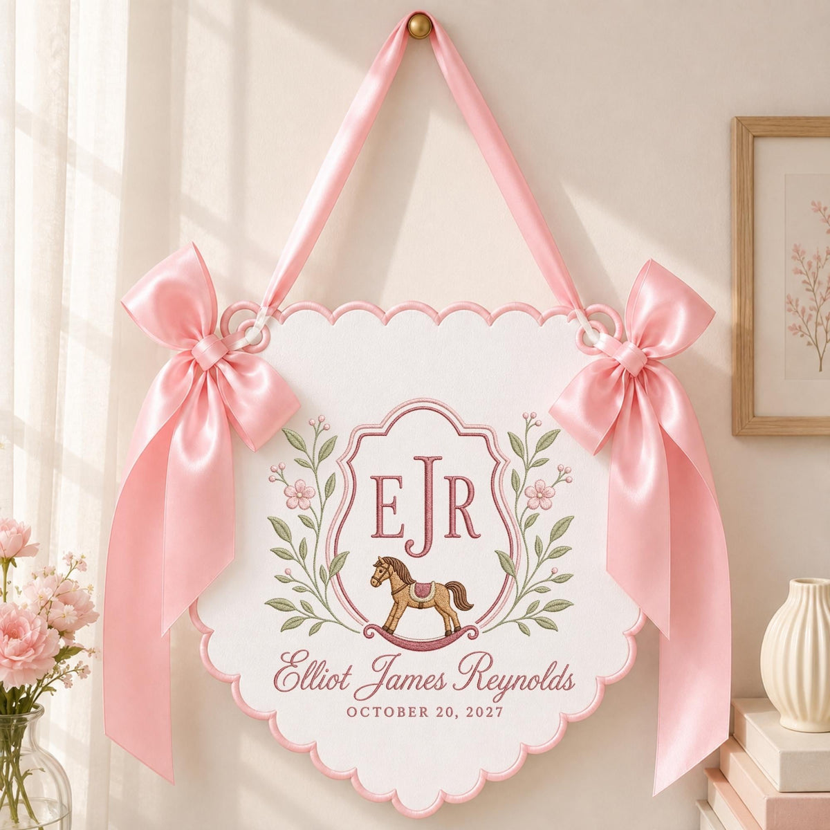May include: A white scalloped wall hanging sign featuring a pink satin ribbon for suspension and two large pink bows on the sides. The center displays the initials EJR, a brown rocking horse illustration, and the text Elliot James Reynolds October 20, 2027.