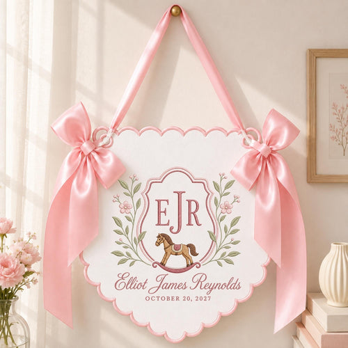 May include: A white scalloped wall hanging sign featuring a pink satin ribbon for suspension and two large pink bows on the sides. The center displays the initials EJR, a brown rocking horse illustration, and the text Elliot James Reynolds October 20, 2027.