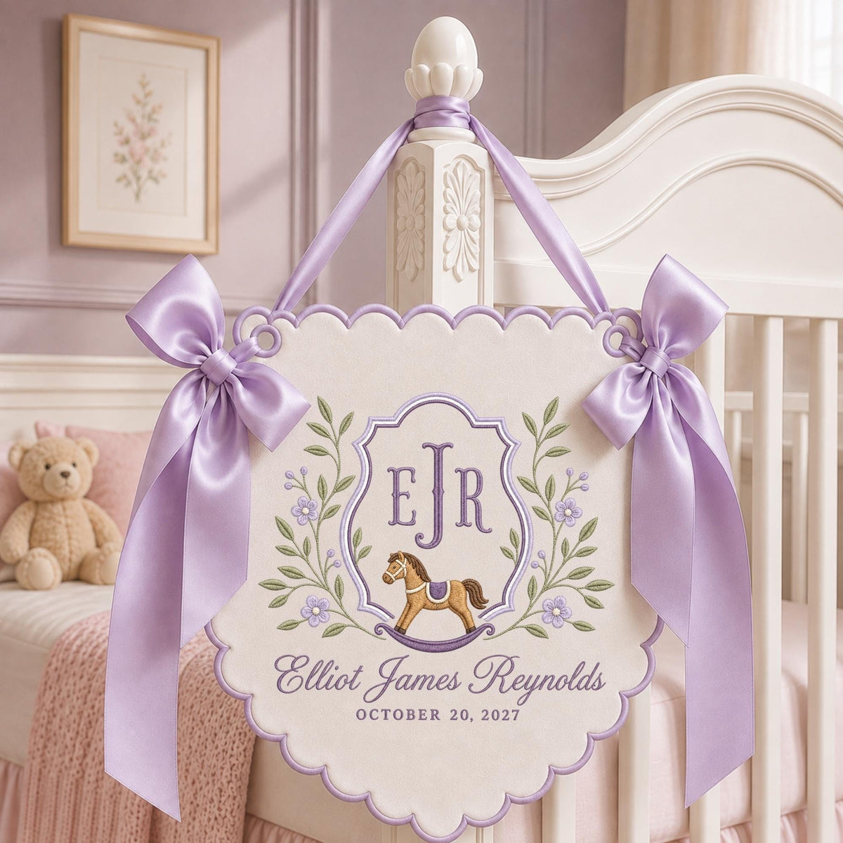 May include: A cream-colored scalloped wall hanging featuring an embroidered purple crest with the initials EJR, a brown rocking horse, and green floral vines. The text reads Elliot James Reynolds and OCTOBER 20, 2027, accented by two lavender satin bows.