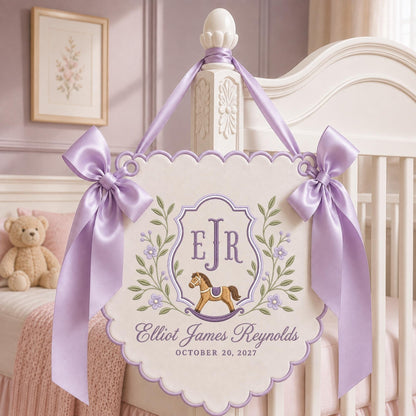 May include: A cream-colored scalloped wall hanging featuring an embroidered purple crest with the initials EJR, a brown rocking horse, and green floral vines. The text reads Elliot James Reynolds and OCTOBER 20, 2027, accented by two lavender satin bows.