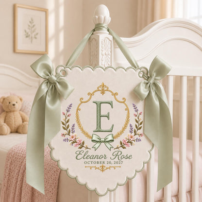 May include: A cream-colored fabric wall hanging with scalloped edges, featuring an embroidered gold frame, a large green letter E, and floral accents. It displays the name Eleanor Rose and the date OCTOBER 20, 2027, with sage green satin bows.