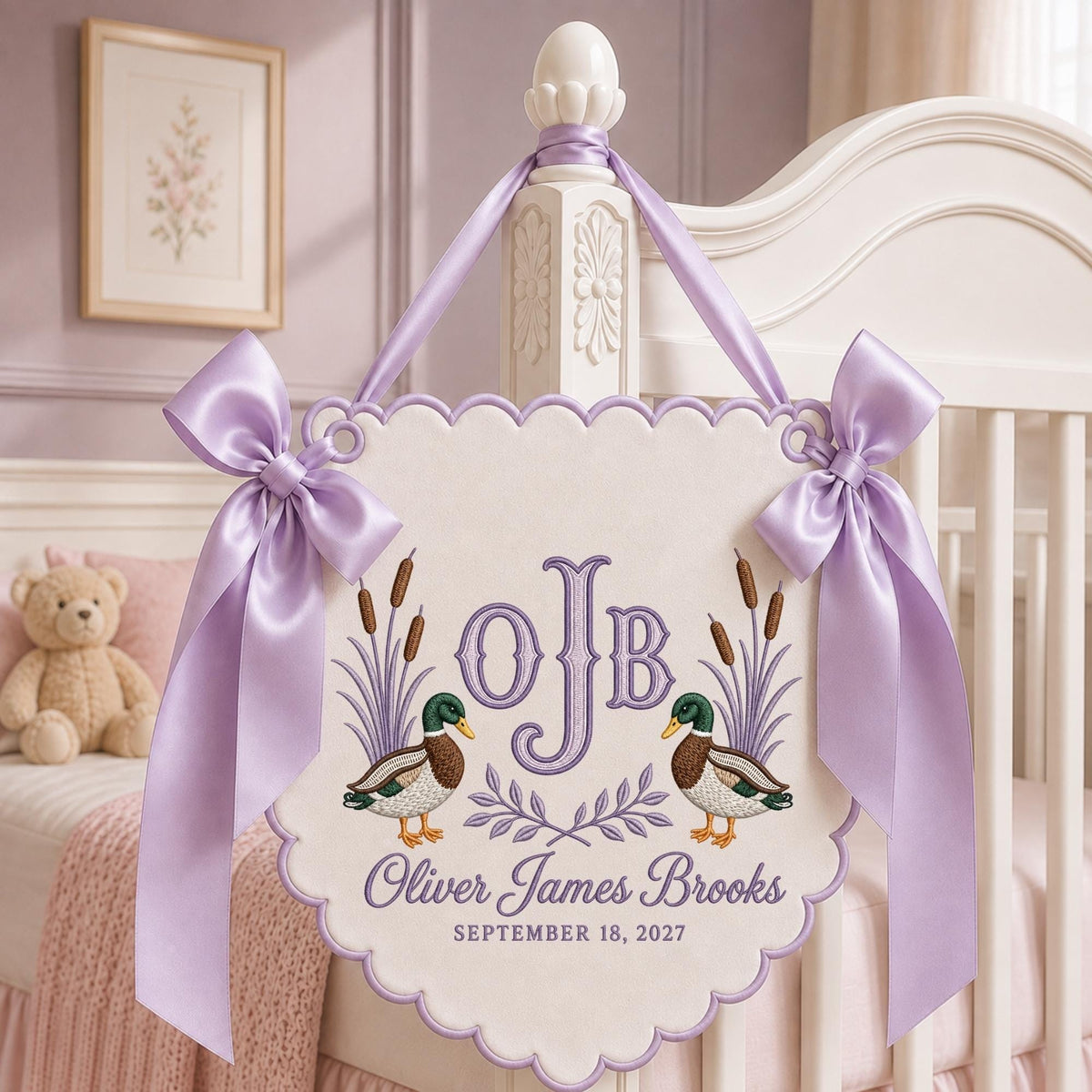 May include: A scalloped, off-white fabric wall hanging featuring an embroidered monogram OJB, two mallard ducks, and cattails. The text reads Oliver James Brooks and SEPTEMBER 18, 2027. It is decorated with two lavender satin bows and a matching ribbon.