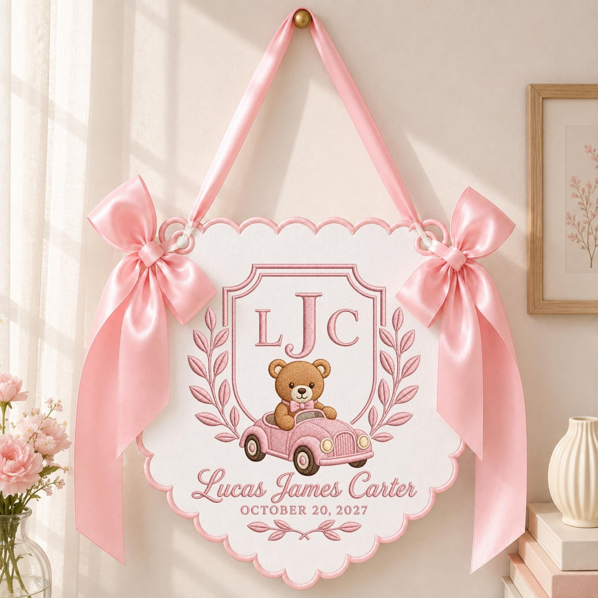 May include: A white scalloped wall hanging featuring a brown teddy bear in a pink car, embroidered with the initials LJC, the name Lucas James Carter, and the date October 20, 2027. The sign is decorated with pink satin bows and a pink ribbon hanger.