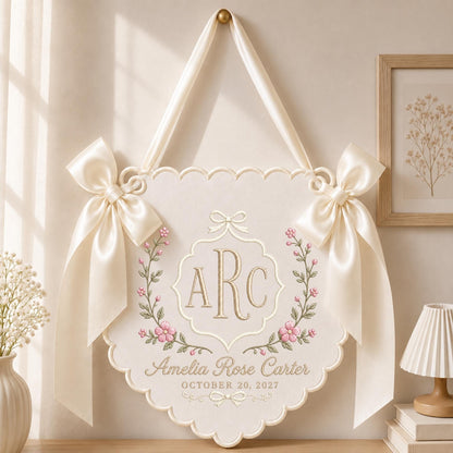 May include: A cream-colored fabric wall hanging with a scalloped edge, featuring two large satin bows on the sides and a ribbon for hanging. The center displays the embroidered monogram ARC, the name Amelia Rose Carter, and October 20, 2027.