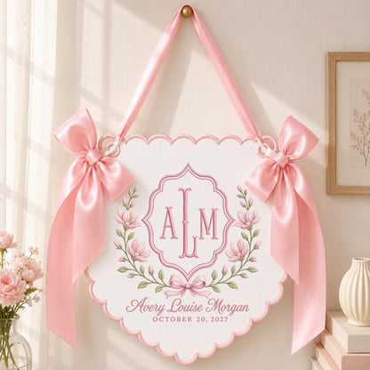 May include: A white scalloped wall hanging featuring a pink embroidered monogram ALM, floral wreath, and the text Avery Louise Morgan October 20, 2027. The sign is suspended by a pink satin ribbon and decorated with two large pink satin bows on each side.
