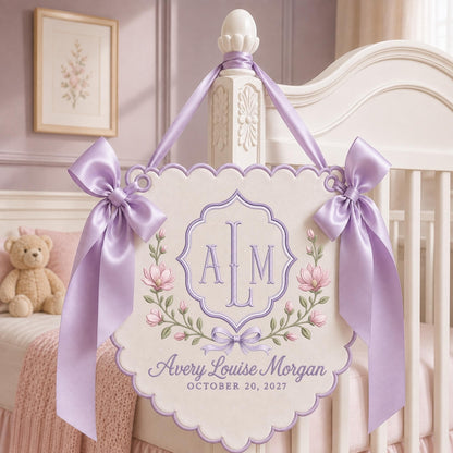 May include: A cream-colored fabric wall hanging with scalloped edges, featuring lavender satin bows on each side. The center displays an embroidered monogram ALM, floral vines, and the text Avery Louise Morgan and OCTOBER 20, 2027 in lavender.