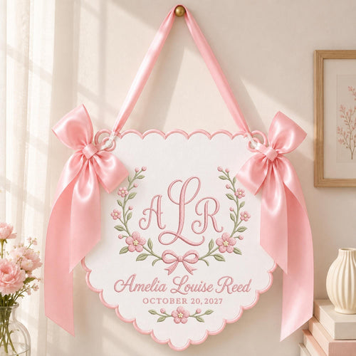May include: A white scalloped wall hanging featuring pink embroidered floral wreaths, a central monogram with the letters ALR, and the text Amelia Louise Reed OCTOBER 20, 2027. The sign is suspended by a pink satin ribbon with large side bows.