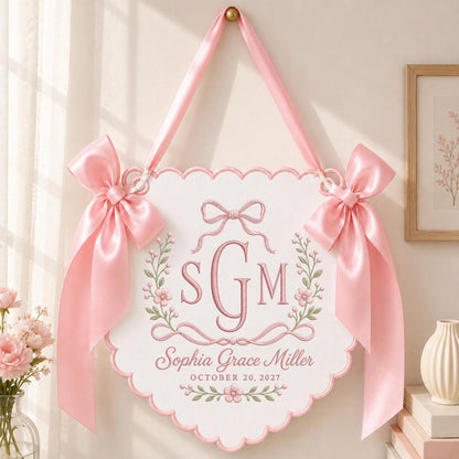 May include: A white scalloped wall hanging featuring pink embroidered floral accents and a central monogram with the letters SGM. Below the monogram, the text reads Sophia Grace Miller and OCTOBER 20, 2027. Two large pink satin bows adorn the top corners.