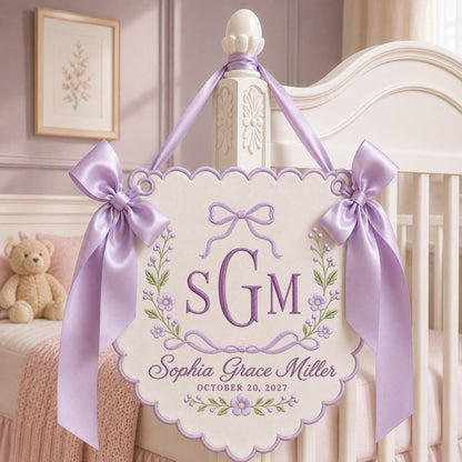 May include: A cream-colored scalloped wall hanging featuring purple embroidery of the monogram SGM, the name Sophia Grace Miller, and the date October 20, 2027. The piece is decorated with floral vines and bows, finished with two large lavender satin ribbons.