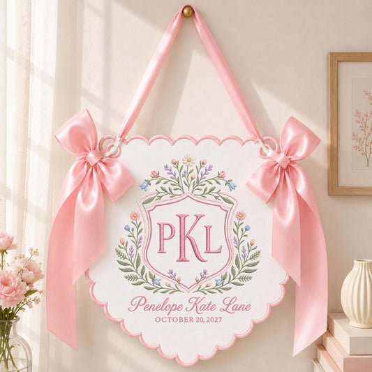 May include: A white scalloped wall hanging featuring a pink embroidered crest with the letters PKL, surrounded by floral motifs. Below the crest, the text reads Penelope Kate Lane and OCTOBER 20, 2027. The sign is accented with two large pink satin bows.