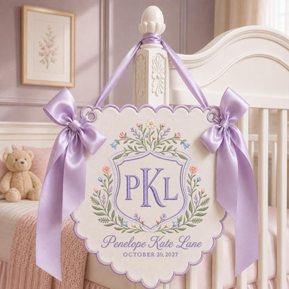 May include: A cream-colored fabric wall hanging with a scalloped edge, featuring purple embroidered initials PKL inside a shield, surrounded by floral motifs. It includes the name Penelope Kate Lane and the date OCTOBER 20, 2027, with lavender satin bows.