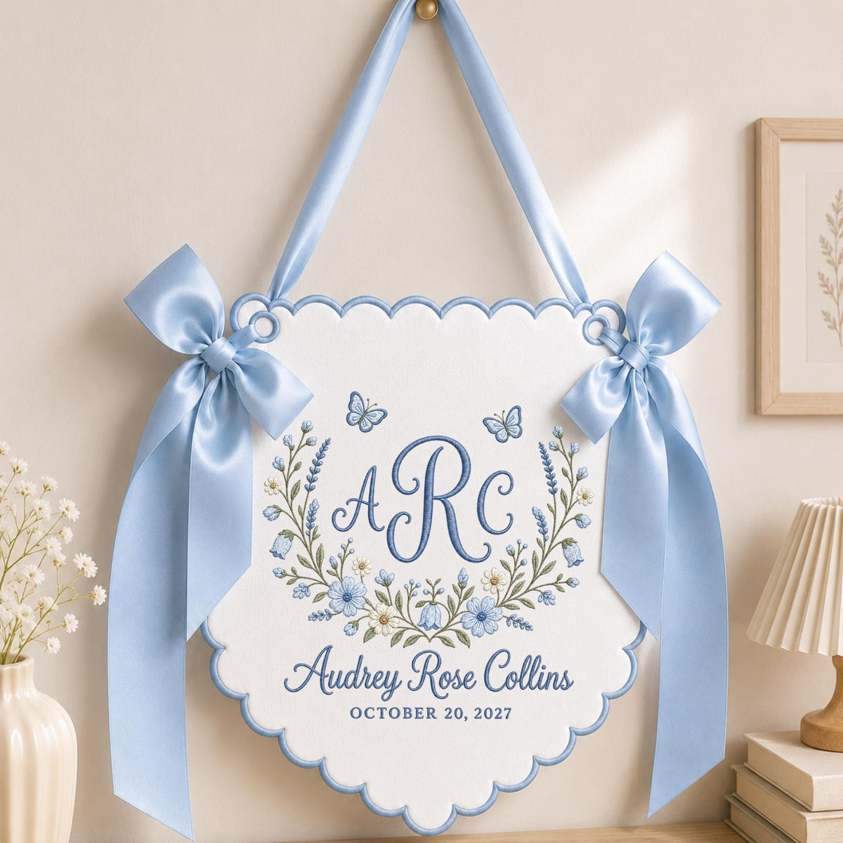 May include: A white scalloped wall hanging banner with light blue satin ribbon bows on each side. The center features blue embroidered floral wreaths, butterflies, the monogram ARC, the name Audrey Rose Collins, and the date OCTOBER 20, 2027.