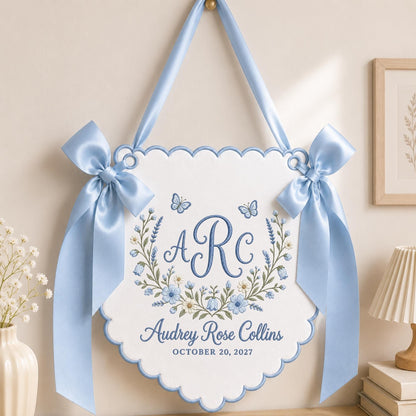 May include: A white scalloped wall hanging banner with light blue satin ribbon bows on each side. The center features blue embroidered floral wreaths, butterflies, the monogram ARC, the name Audrey Rose Collins, and the date OCTOBER 20, 2027.