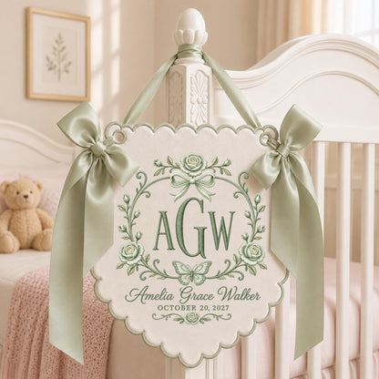 May include: A cream-colored fabric wall hanging with scalloped edges, featuring sage green embroidery of the initials AGW, a butterfly, and roses. The text reads Amelia Grace Walker and OCTOBER 20, 2027. It is adorned with two sage green satin ribbons.