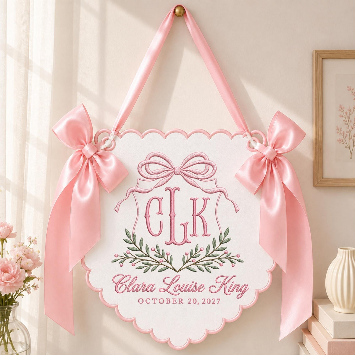 May include: A white scalloped wall hanging featuring pink embroidered details, including a bow, a monogram with the letters CLK, and the text Clara Louise King and OCTOBER 20, 2027. The piece is accented with two large pink satin bows and a hanging ribbon.