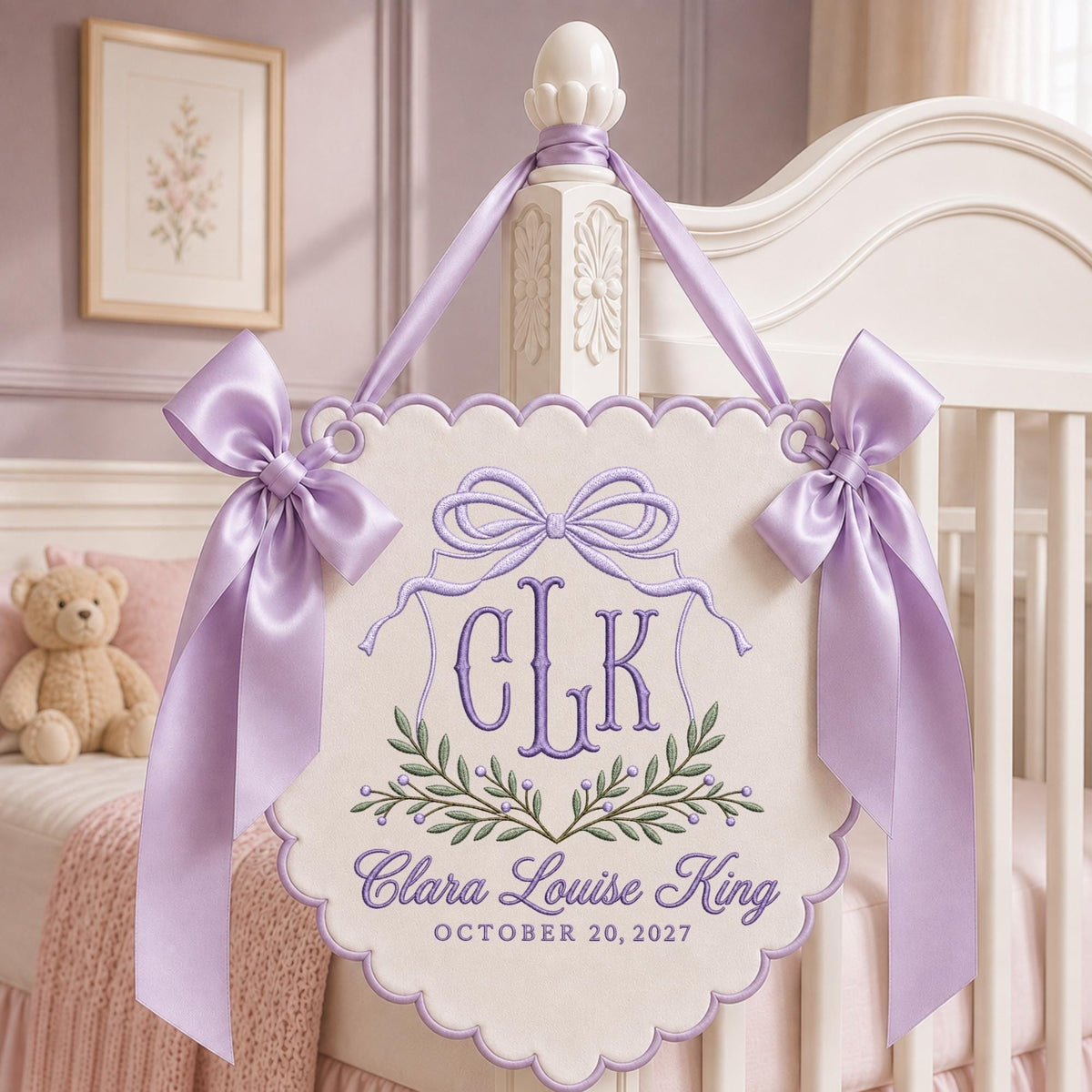 May include: A cream-colored scalloped wall hanging featuring purple embroidery of the monogram CLK, a bow, and a leafy branch. Below, the text reads Clara Louise King and OCTOBER 20, 2027. The piece is accented with two large lavender satin bows.