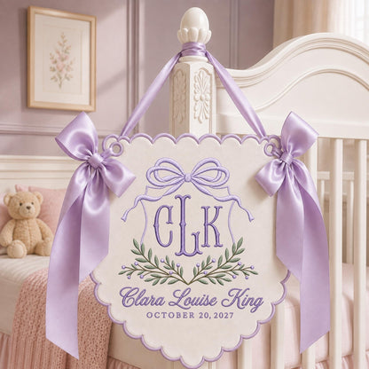 May include: A cream-colored scalloped wall hanging featuring purple embroidery of the monogram CLK, a bow, and a leafy branch. Below, the text reads Clara Louise King and OCTOBER 20, 2027. The piece is accented with two large lavender satin bows.