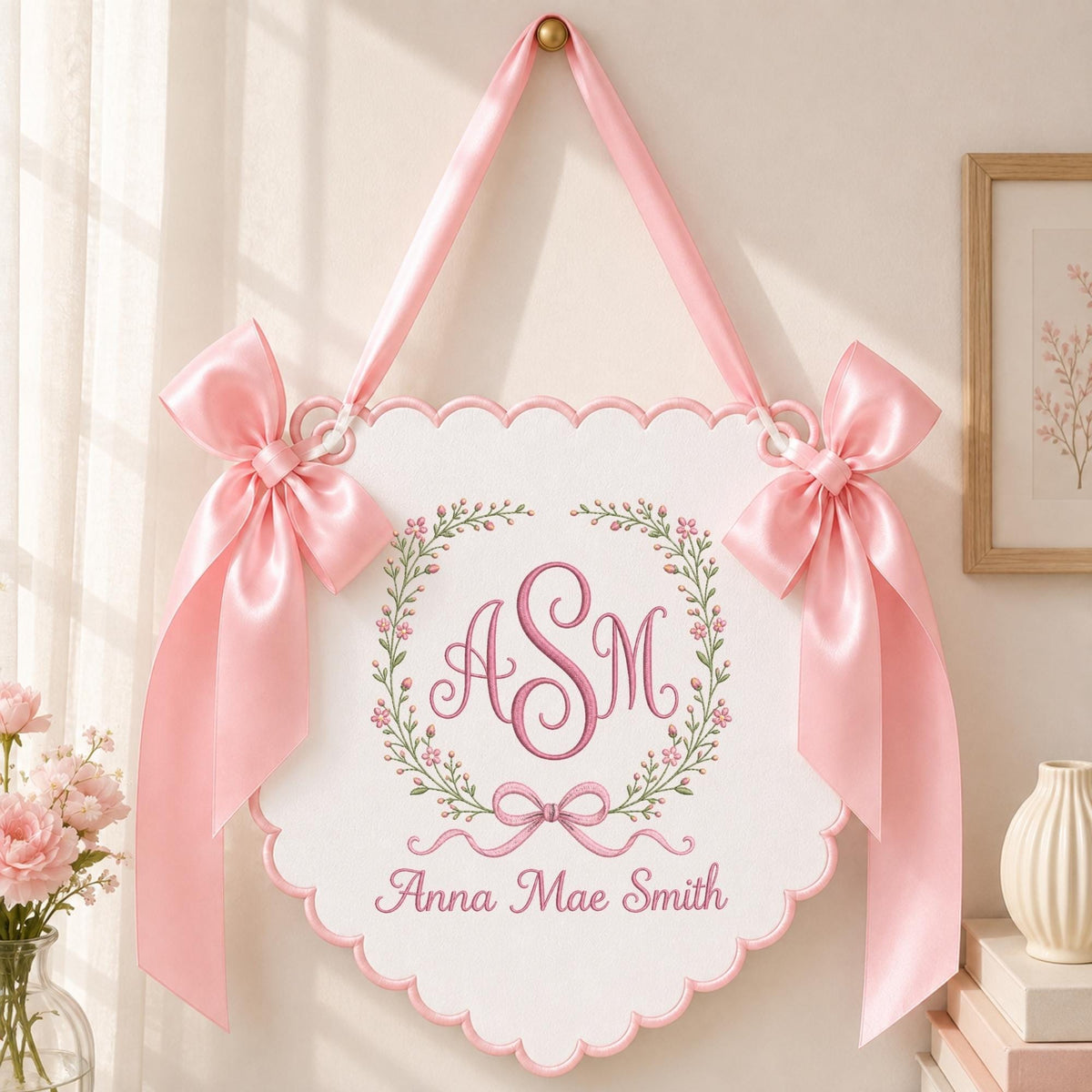 May include: A white scalloped wall hanging featuring a pink embroidered monogram with the letters ASM inside a floral wreath. The sign includes the name Anna Mae Smith in pink text, a pink ribbon hanger, and two large pink satin bows on the sides.