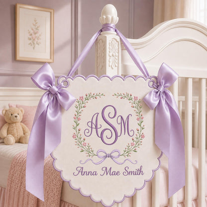 May include: A cream-colored scalloped wall hanging featuring an embroidered floral wreath surrounding the monogram ASM in purple thread. Below the wreath, the name Anna Mae Smith is embroidered in purple. Two lavender satin bows adorn the top corners.