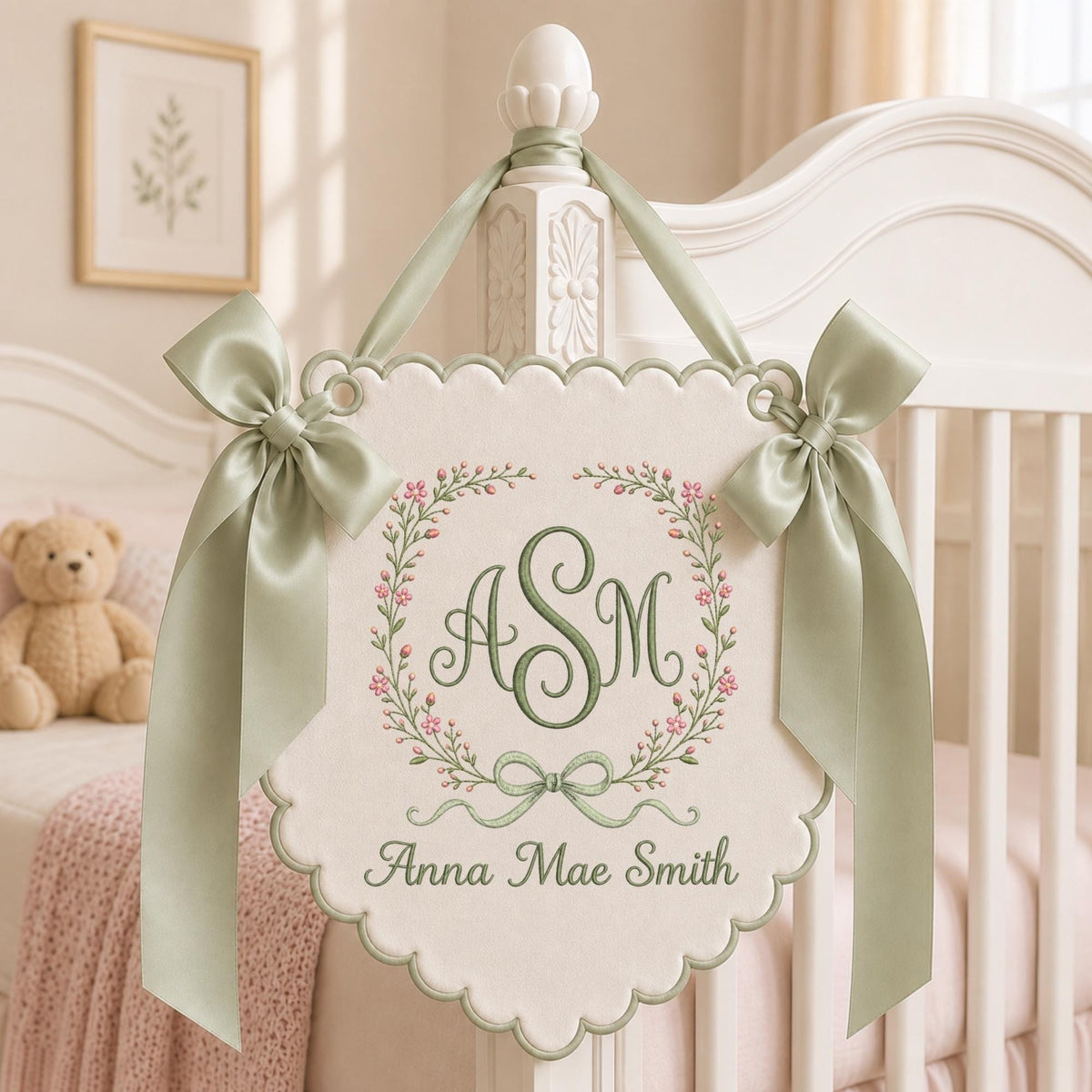 May include: A cream-colored scalloped fabric wall hanging featuring sage green embroidery of the monogram ASM inside a floral wreath with pink buds. The piece is accented with two sage green satin bows and the name Anna Mae Smith in green text.