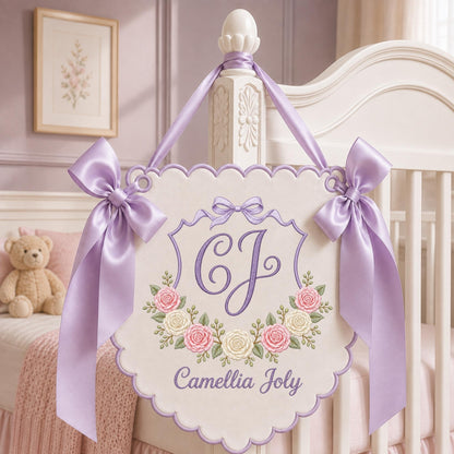 May include: A cream-colored fabric wall hanging with a scalloped edge, featuring an embroidered purple monogram of the letters CJ, a floral wreath of pink and white roses, and the name Camellia Joly in purple script, accented by lavender satin bows.