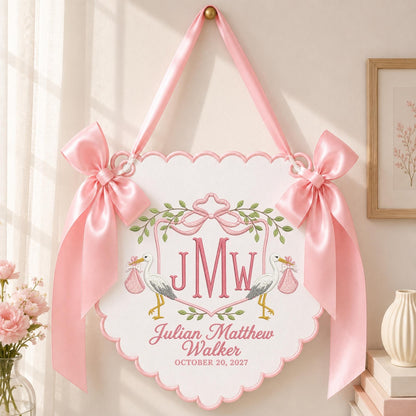 May include: A white scalloped wall hanging featuring pink satin bows and ribbon. The design includes embroidered storks carrying pink bundles, a central monogram JMW, and the text Julian Matthew Walker and the date OCTOBER 20, 2027.