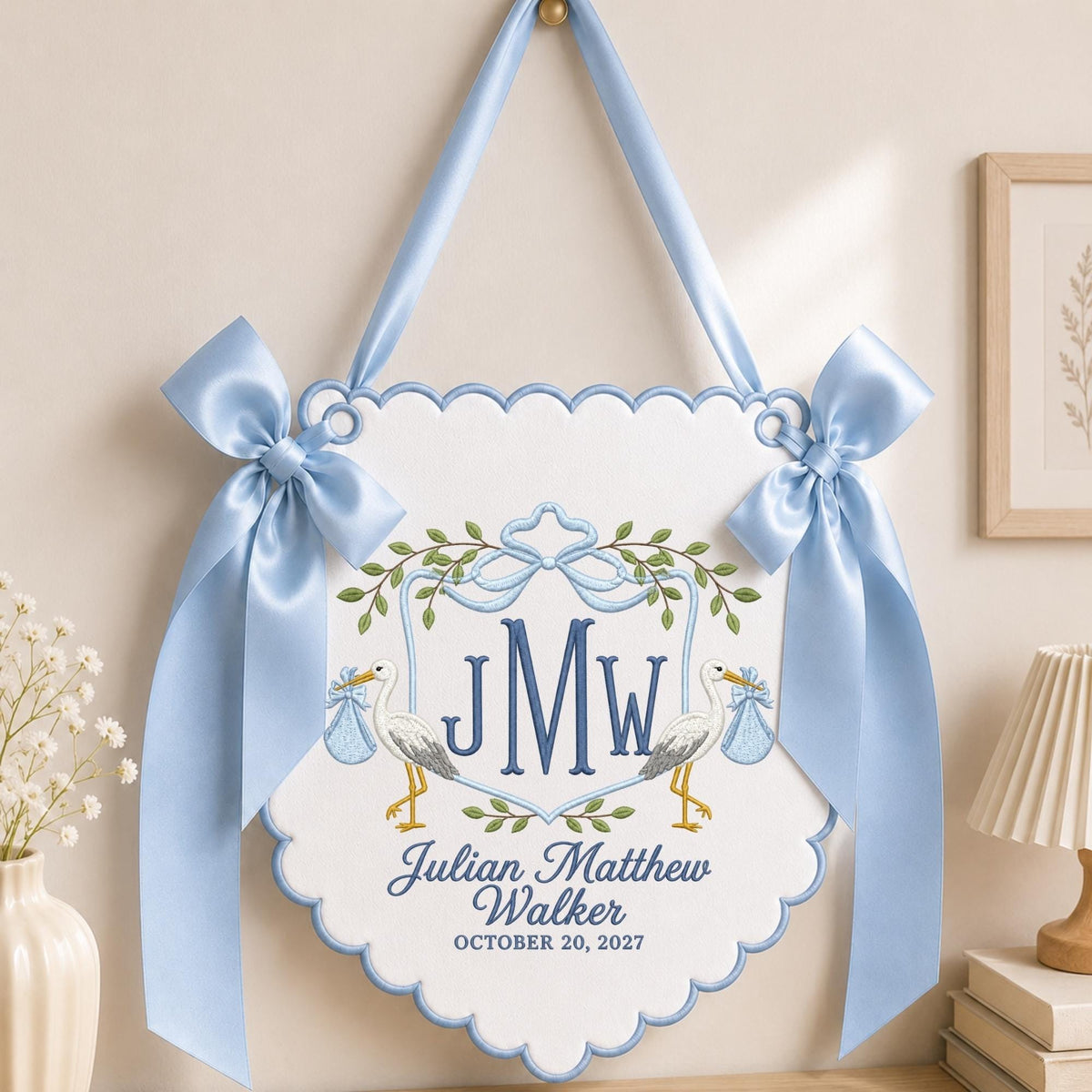 May include: A white scalloped wall hanging featuring blue embroidery of two storks carrying blue bundles, a central monogram JMW, and the text Julian Matthew Walker OCTOBER 20, 2027. The piece is accented with two large light blue satin bows.