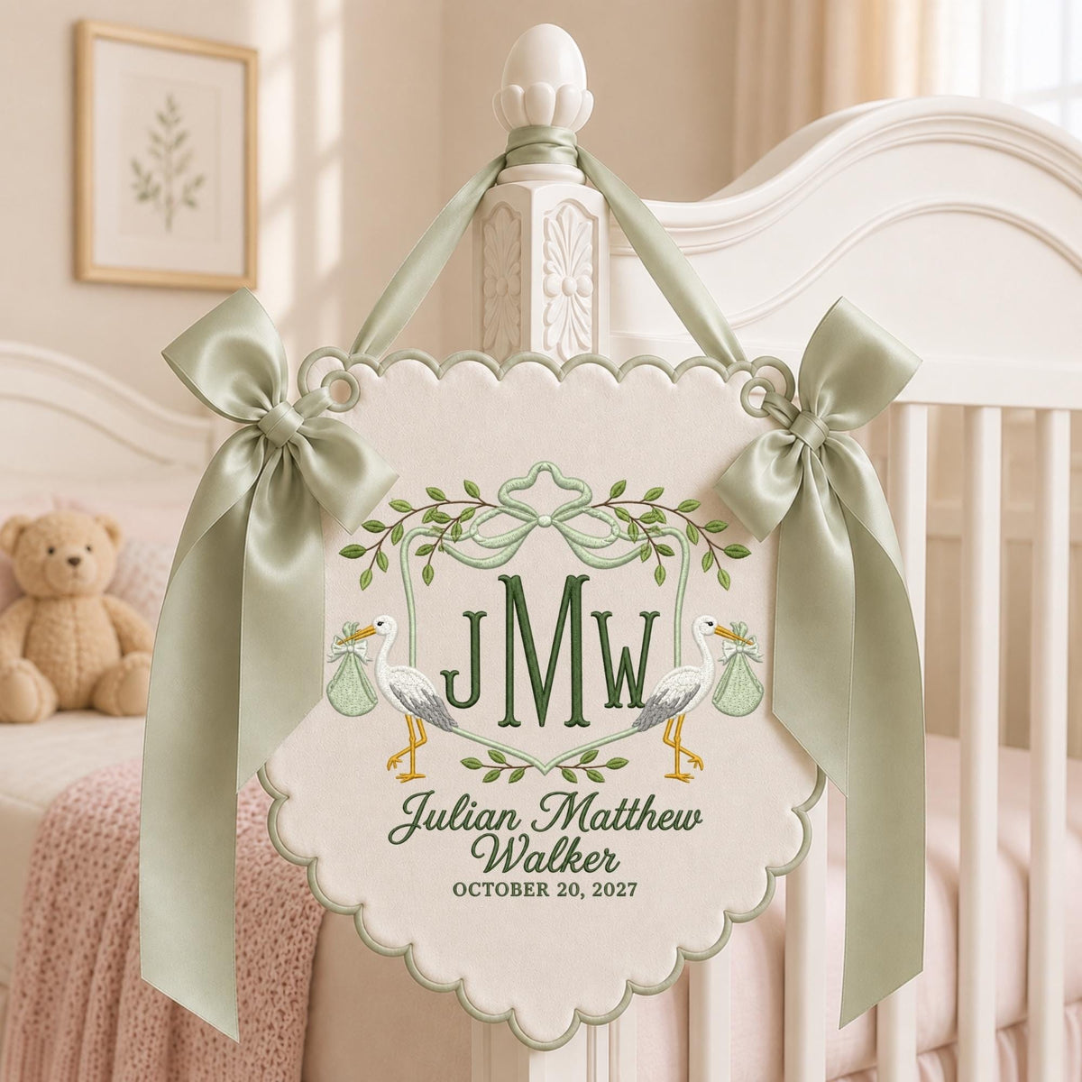 May include: A cream-colored scalloped fabric wall hanging featuring embroidered green storks, leafy vines, and a central monogram JMW. The text reads Julian Matthew Walker and October 20, 2027. It is adorned with two sage green satin ribbons.