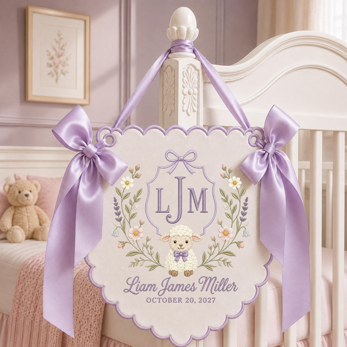 May include: A scalloped, cream-colored fabric wall hanging featuring a central monogram LJM, a small lamb illustration, and the text Liam James Miller October 20, 2027. The banner is decorated with floral embroidery and two lavender satin bows.