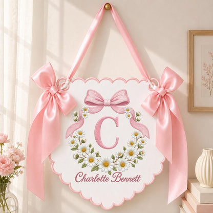 May include: A white scalloped wall hanging featuring a pink embroidered letter C surrounded by a daisy wreath and the name Charlotte Bennett. The sign is accented with two large pink satin bows on the sides and a pink ribbon for hanging.