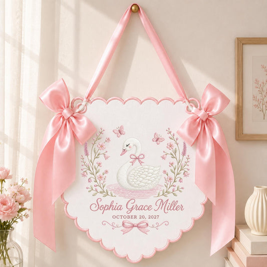 May include: A white scalloped wall hanging featuring a central illustration of a white swan with a pink bow, surrounded by floral vines and butterflies. The text reads Sophia Grace Miller and OCTOBER 20, 2027. It is suspended by pink satin ribbons.