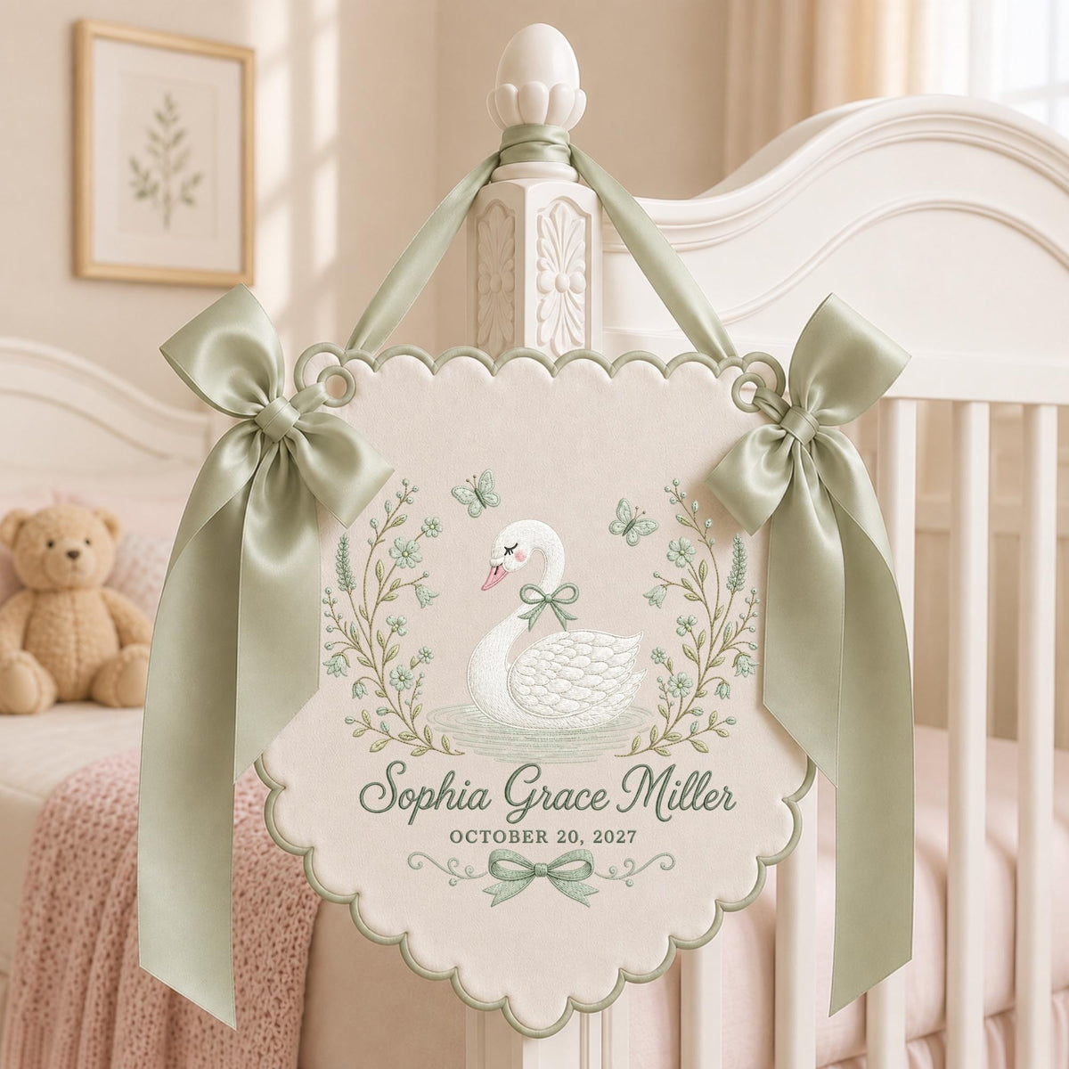 May include: A cream-colored scalloped wall hanging featuring a white swan illustration, sage green floral accents, and butterflies. The text reads Sophia Grace Miller and OCTOBER 20, 2027. It is decorated with two sage green satin bows and a ribbon hanger.
