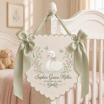 May include: A cream-colored scalloped wall hanging featuring a white swan illustration, sage green floral accents, and butterflies. The text reads Sophia Grace Miller and OCTOBER 20, 2027. It is decorated with two sage green satin bows and a ribbon hanger.