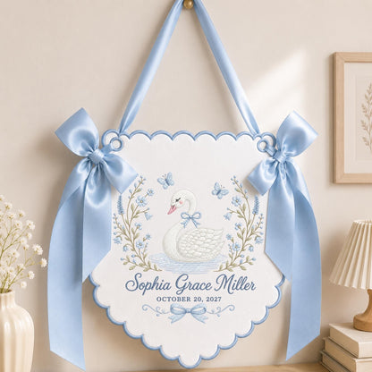 May include: A white scalloped wall hanging featuring an embroidered white swan, blue floral wreaths, and butterflies. The text reads Sophia Grace Miller and OCTOBER 20, 2027. It is accented with large light blue satin bows and a matching hanging ribbon.