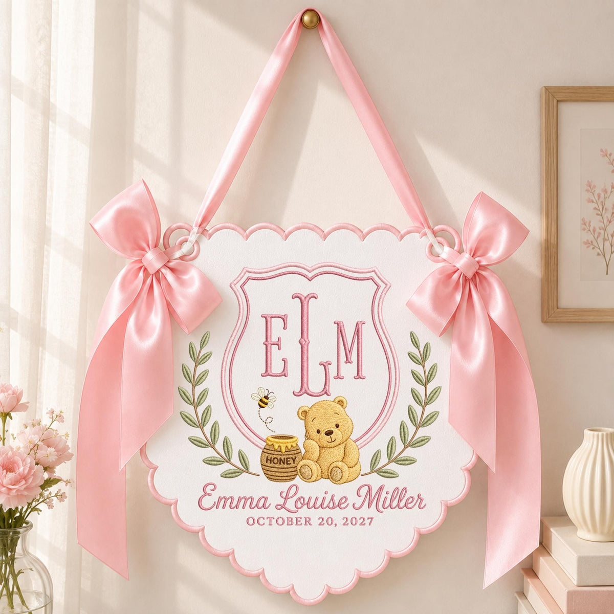 May include: A white scalloped wall hanging featuring a pink embroidered monogram ELM, a teddy bear with a honey pot, and the text Emma Louise Miller OCTOBER 20, 2027. The sign is decorated with green leaf wreaths and two large pink satin bows.
