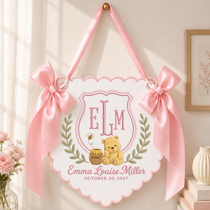 May include: A white scalloped wall hanging featuring a pink embroidered monogram ELM, a teddy bear with a honey pot, and the text Emma Louise Miller OCTOBER 20, 2027. The sign is decorated with green leaf wreaths and two large pink satin bows.