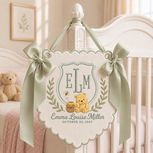 May include: A cream-colored fabric wall hanging with scalloped edges, featuring sage green satin bows on each side. The embroidery includes the monogram ELM, a yellow bear, a honey pot, a bee, and the text Emma Louise Miller October 20, 2027.