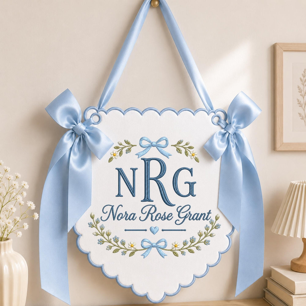 May include: A white scalloped wall hanging banner featuring blue embroidered text reading NRG and Nora Rose Grant, accented with floral motifs and blue bows. The banner is suspended by a matching light blue satin ribbon for display.