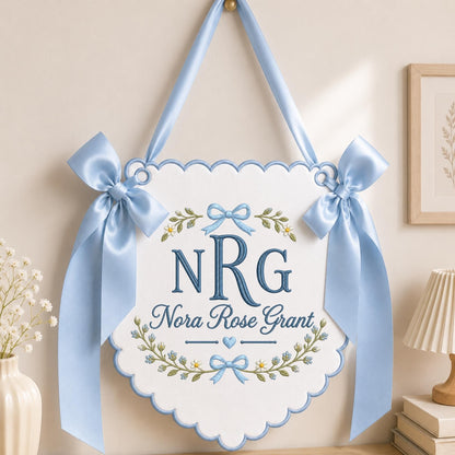 May include: A white scalloped wall hanging banner featuring blue embroidered text reading NRG and Nora Rose Grant, accented with floral motifs and blue bows. The banner is suspended by a matching light blue satin ribbon for display.
