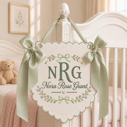May include: A cream-colored fabric wall hanging with scalloped edges, featuring sage green embroidery of the initials NRG and the name Nora Rose Grant. The banner is decorated with floral motifs and bows, and is suspended by sage green satin ribbons.