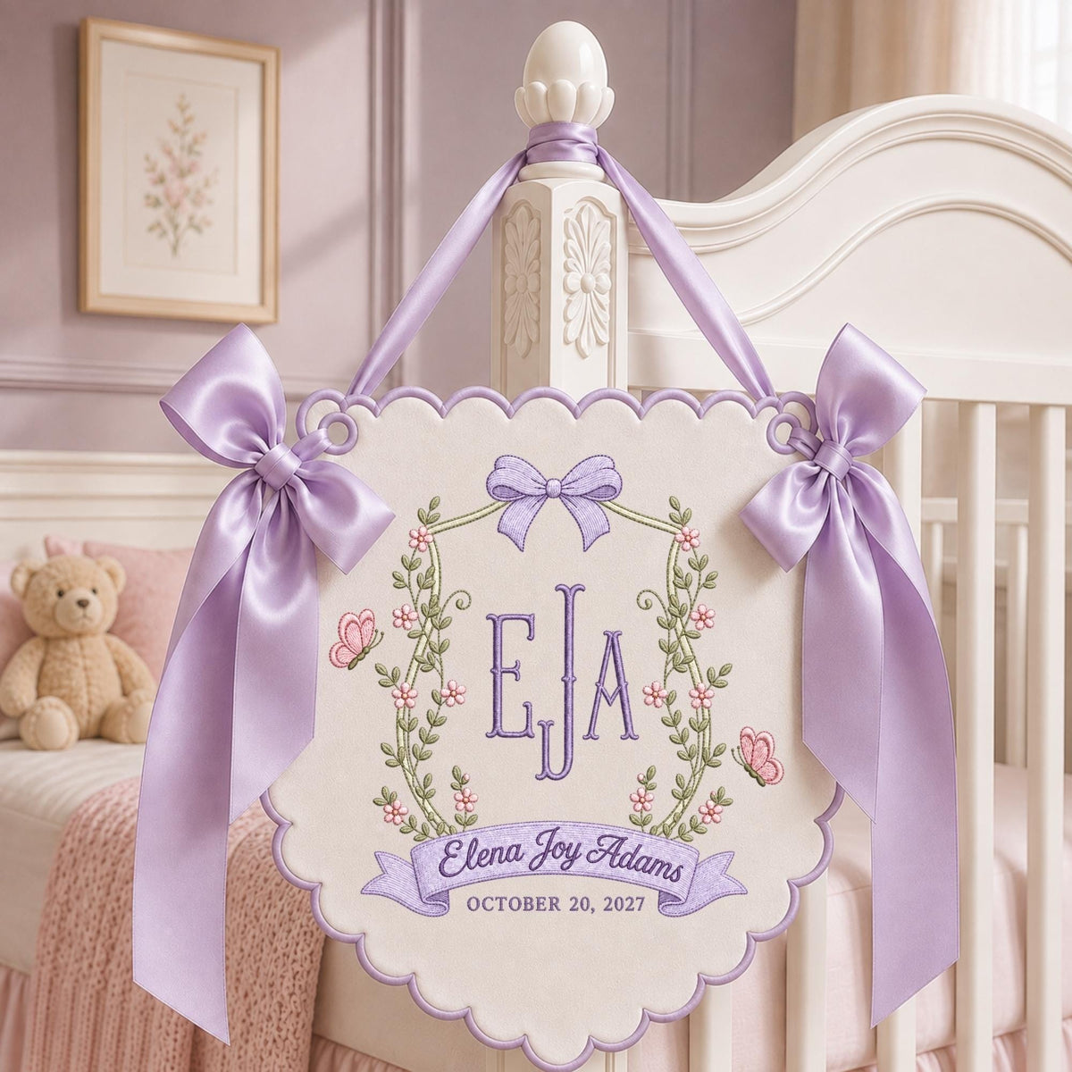 May include: A cream-colored fabric wall hanging with scalloped edges, featuring purple embroidery of the initials EJA, a floral wreath, butterflies, and a bow. It includes the text Elena Joy Adams and October 20, 2027, with lavender satin ribbons.