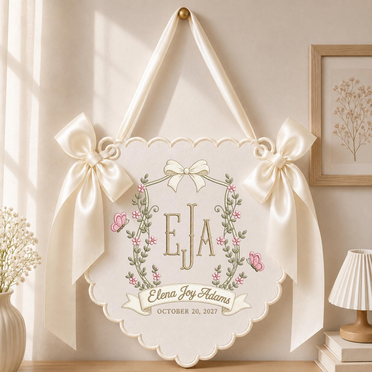May include: A cream-colored fabric wall hanging with scalloped edges, adorned with two large satin bows. The center features an embroidered crest with the initials EJA, pink flowers, green vines, and butterflies, plus the name Elena Joy Adams.